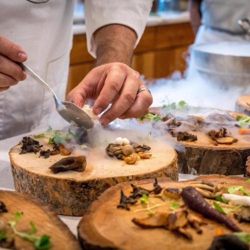 A chef is carefully placing gourmet mushrooms on top of a slice of a tree log which is being used as a plate, dry ice fog is swirling around the chefs hands in a dramatic scene.